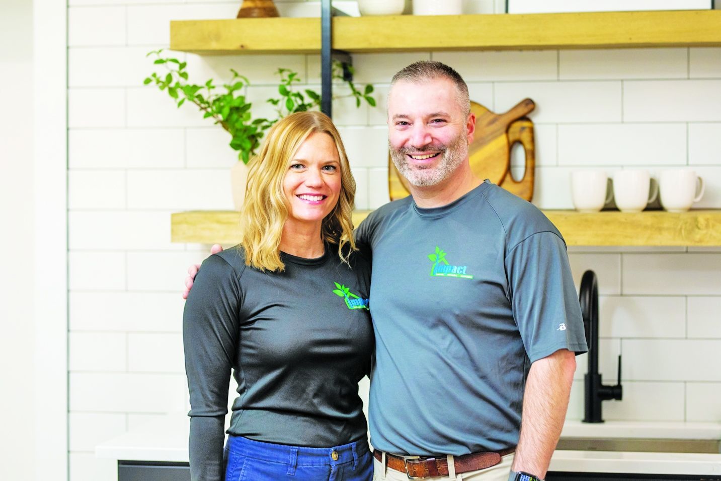 A smiling couple in matching dark gray shirts stand side-by-side in a modern kitchen with wooden shelves and white tile.