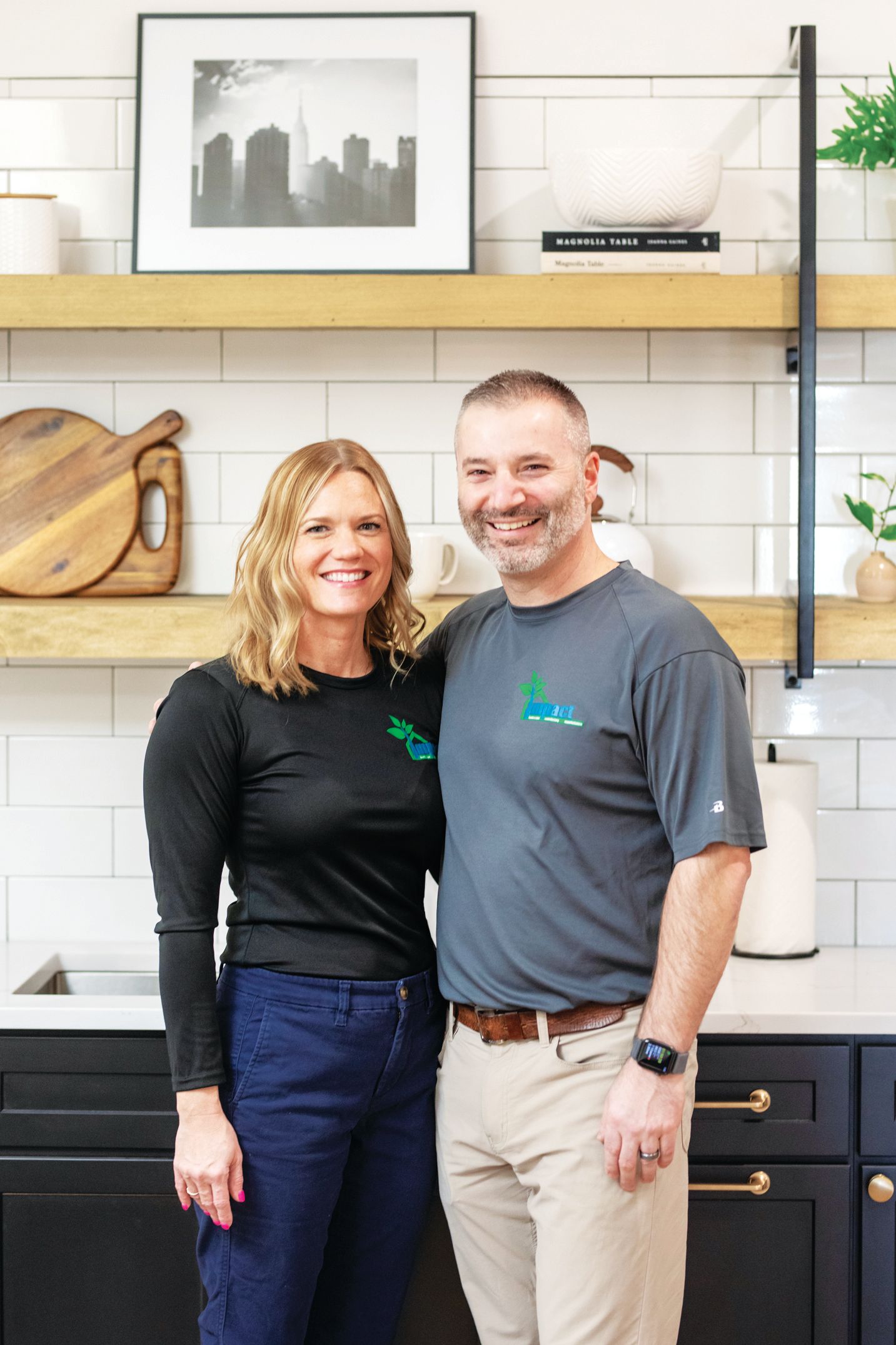 A smiling couple stands side-by-side in a modern kitchen with navy cabinets, white subway tiles, and wooden shelves.