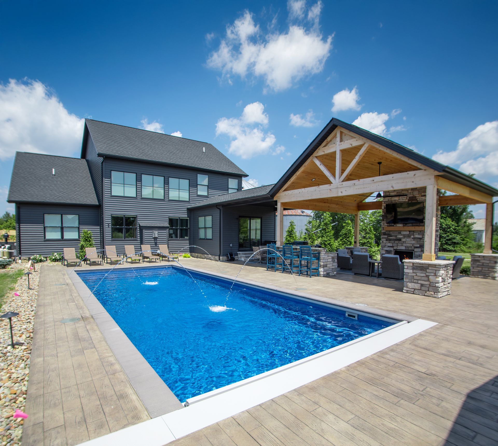 A backyard featuring a rectangular swimming pool next to a dark gray house and a wooden covered outdoor lounge area.