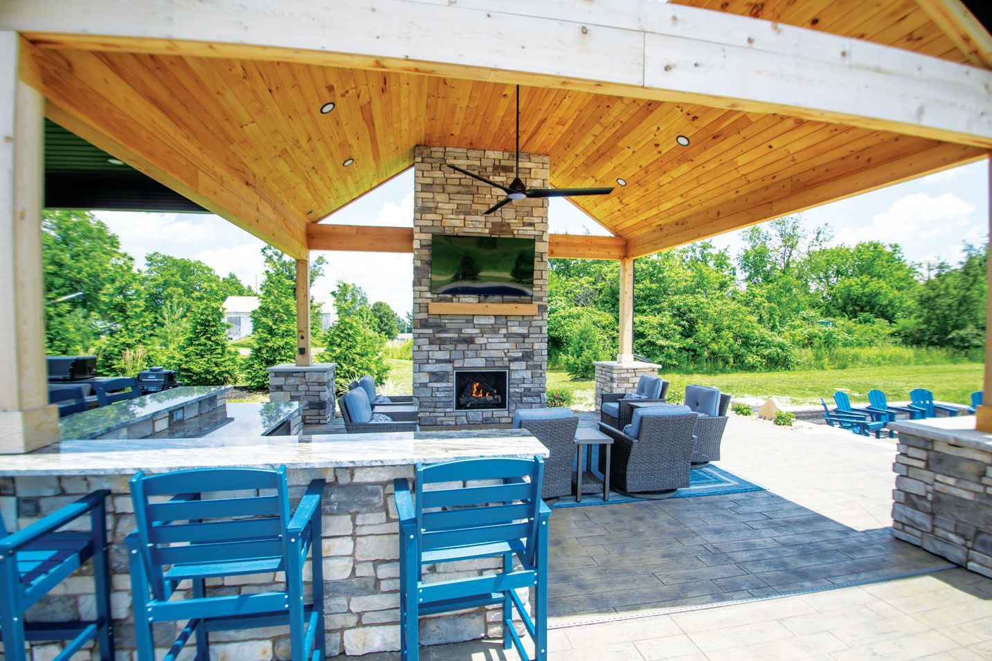 Outdoor patio with a stone fireplace, TV, and wooden ceiling, featuring teal bar stools and blue chairs on a stone floor.