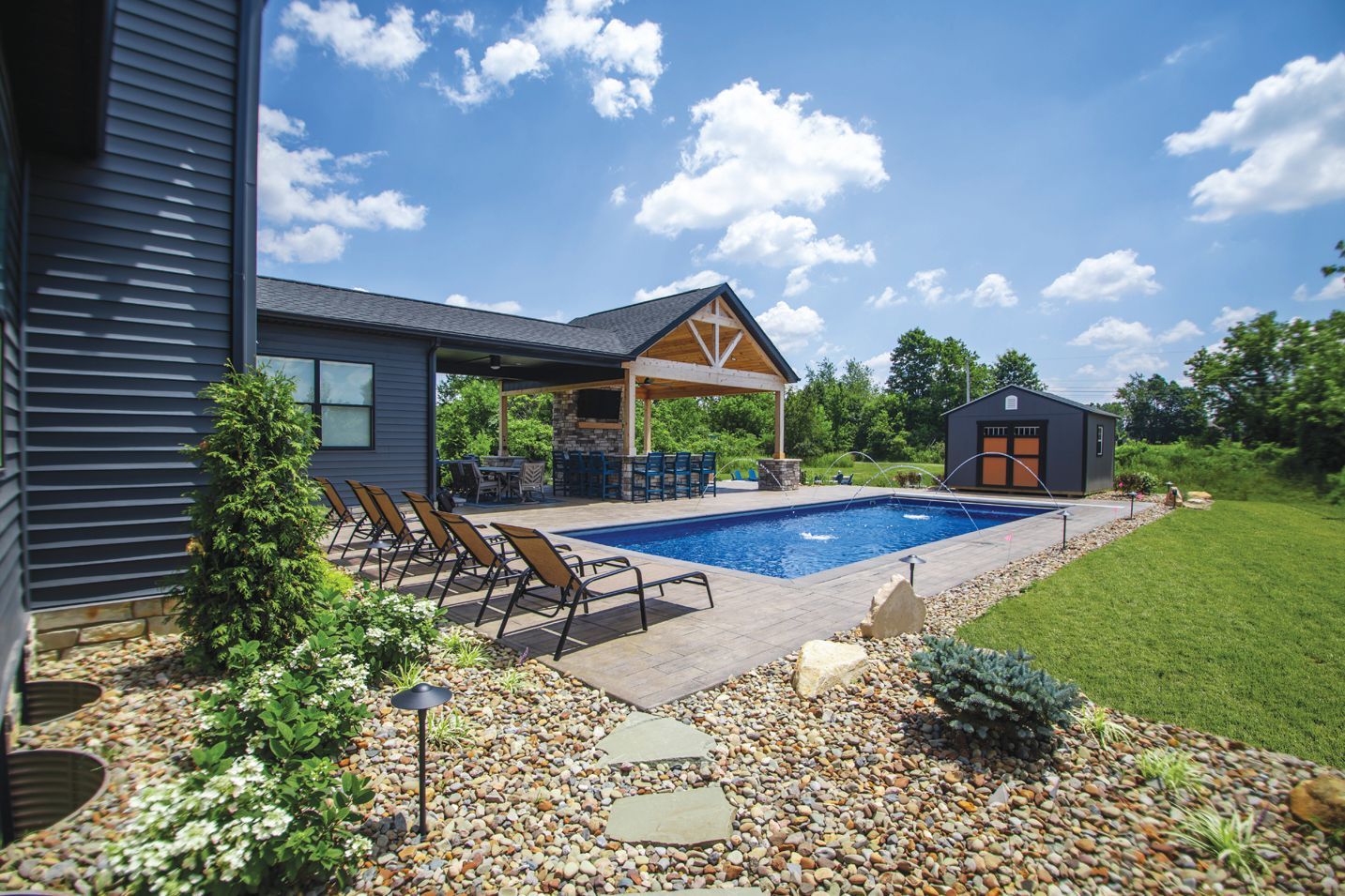 A sunny backyard features a blue swimming pool, several lounge chairs, a covered patio with a grill, and a small shed.