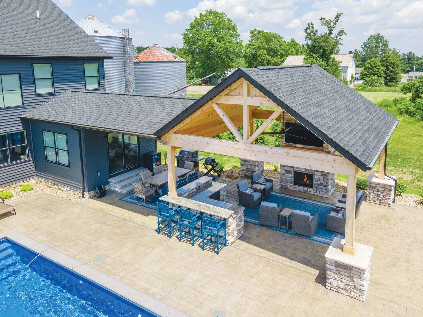 A dark blue house with an attached covered outdoor pavilion featuring a stone fireplace, bar seating, and a pool area.
