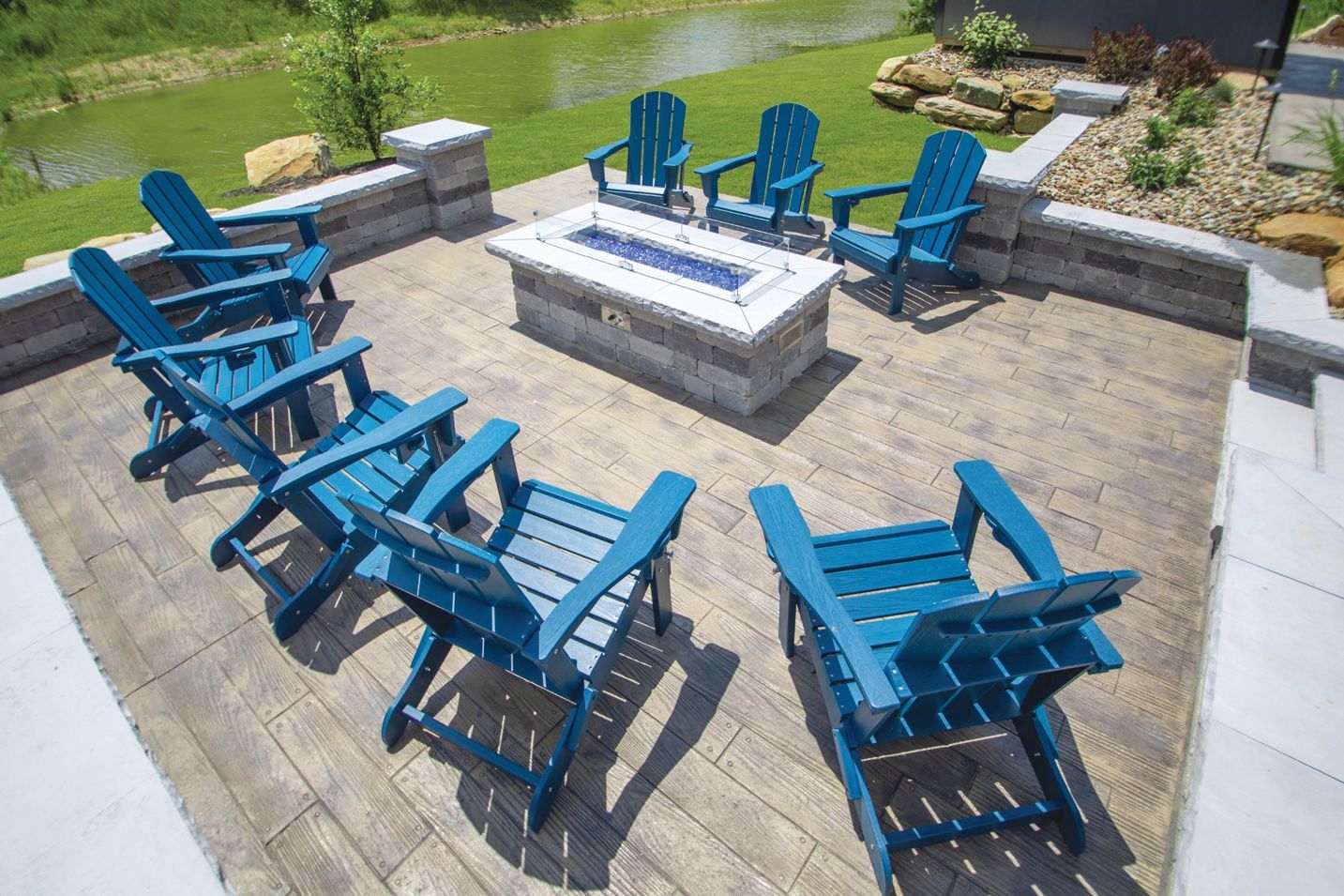 A fire pit with blue Adirondack chairs on a stone patio overlooking a grassy lawn and pond.