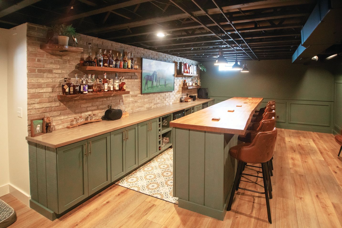 A basement home bar with sage green cabinetry, a wood countertop, brick wall shelving, and brown leather bar stools.