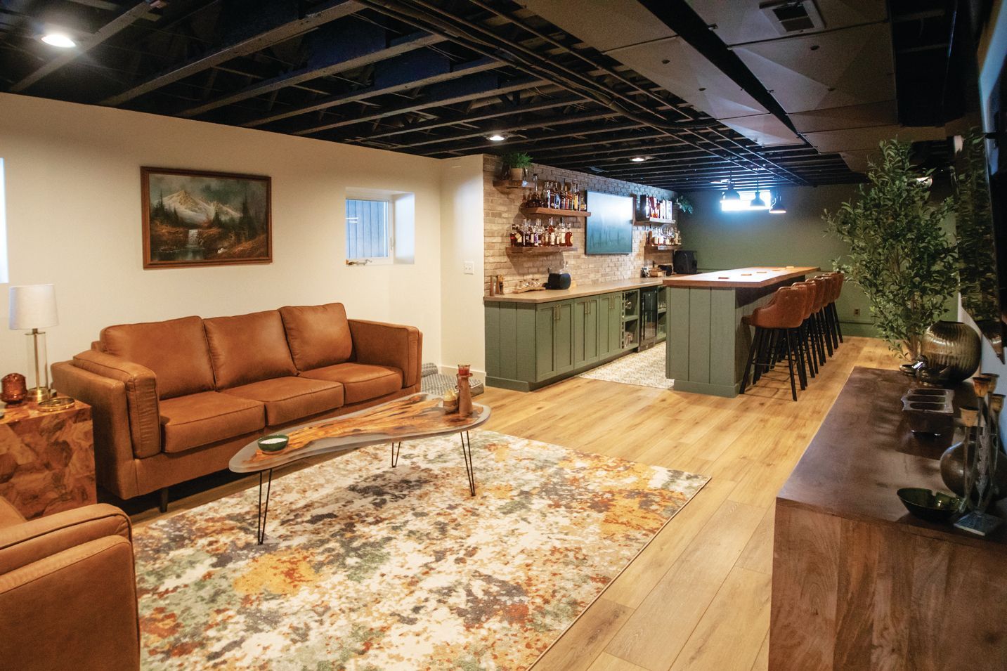 Basement lounge with a leather sofa, patterned rug, stone-accented bar, wood flooring, and an exposed ceiling.