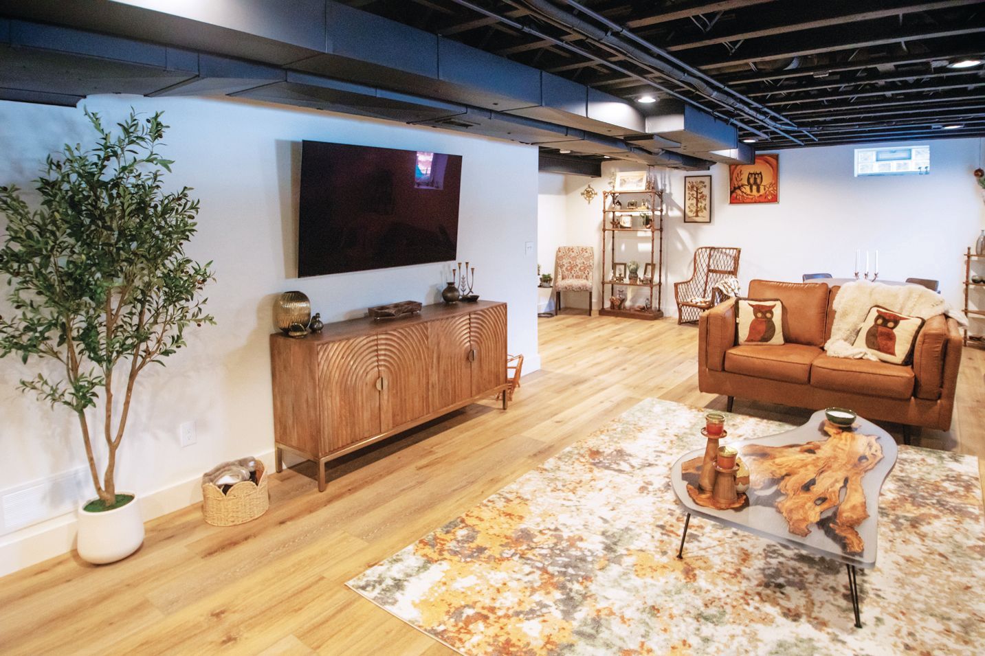 A modern basement living area with light wood floors, a leather sofa, a patterned rug, and a TV mounted above a sideboard.