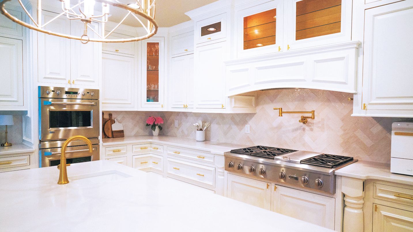 A bright white kitchen with a large island, gold fixtures, a double wall oven, and a gas range with a pot filler.