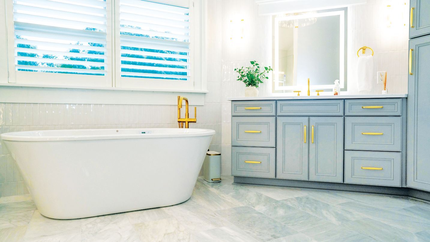 A modern bathroom featuring a white soaking tub, light gray cabinets with gold hardware, and bright lighting.