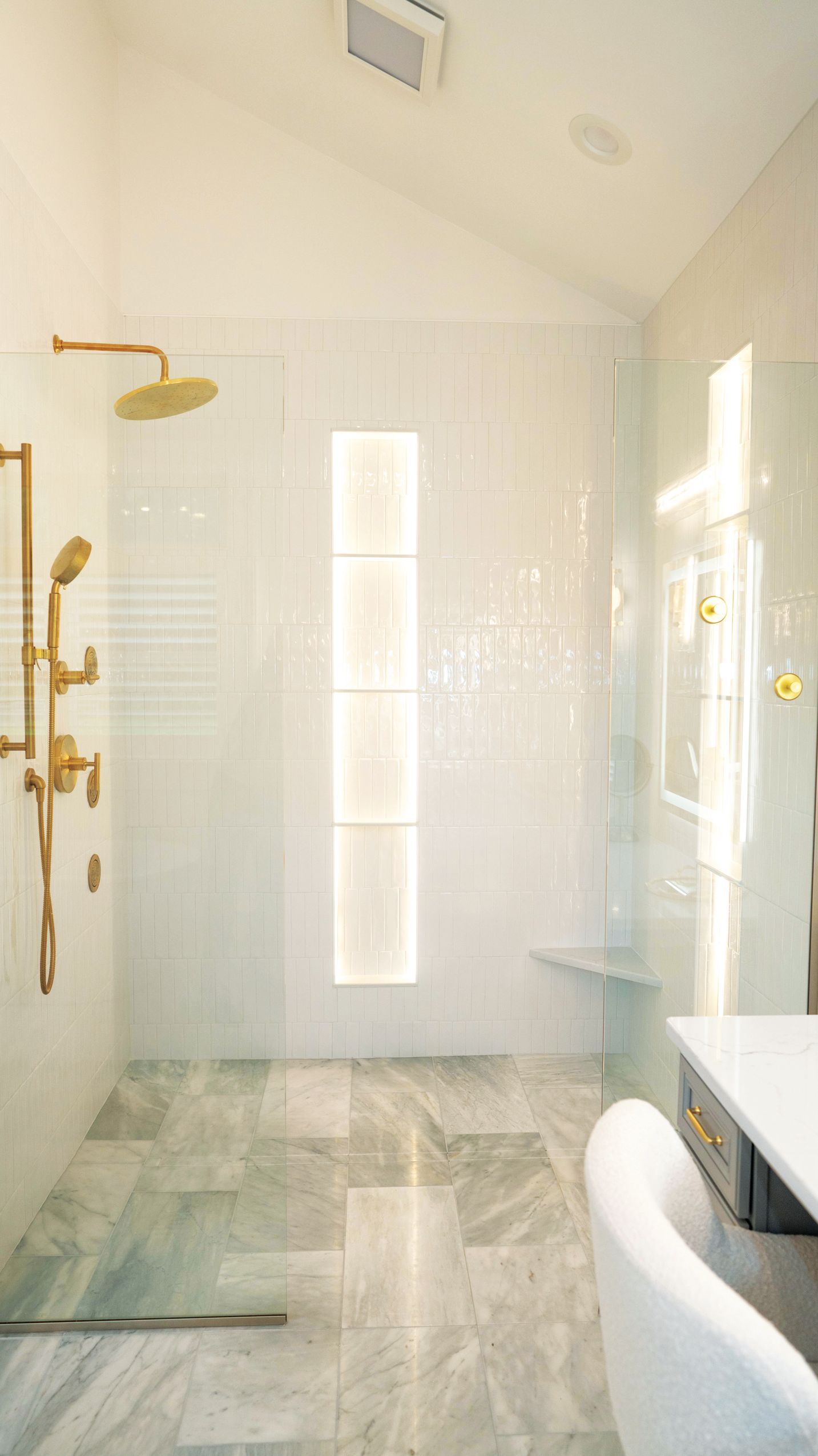 A marble-tiled walk-in shower with gold hardware, a glass partition, a lit vertical wall niche, and a white chair nearby.