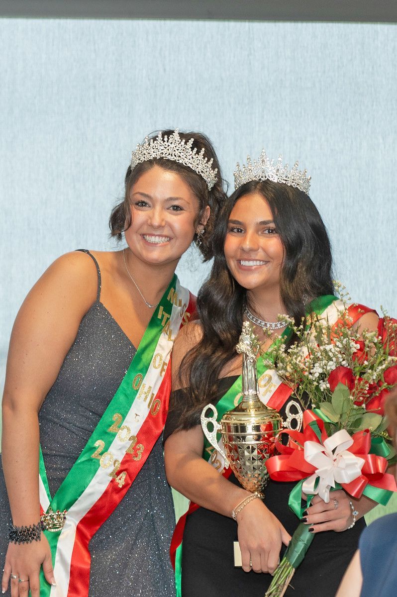 Two people wear tiaras and matching red, white, and green sashes; one holds a golden trophy and bouquet of flowers.