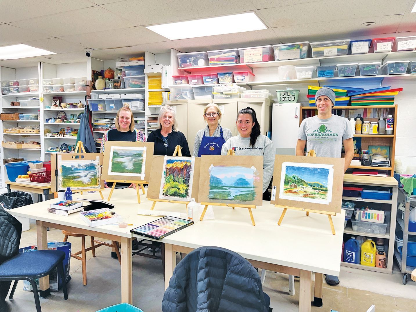 Five people stand behind a row of easels displaying landscape paintings in an art classroom filled with supplies.