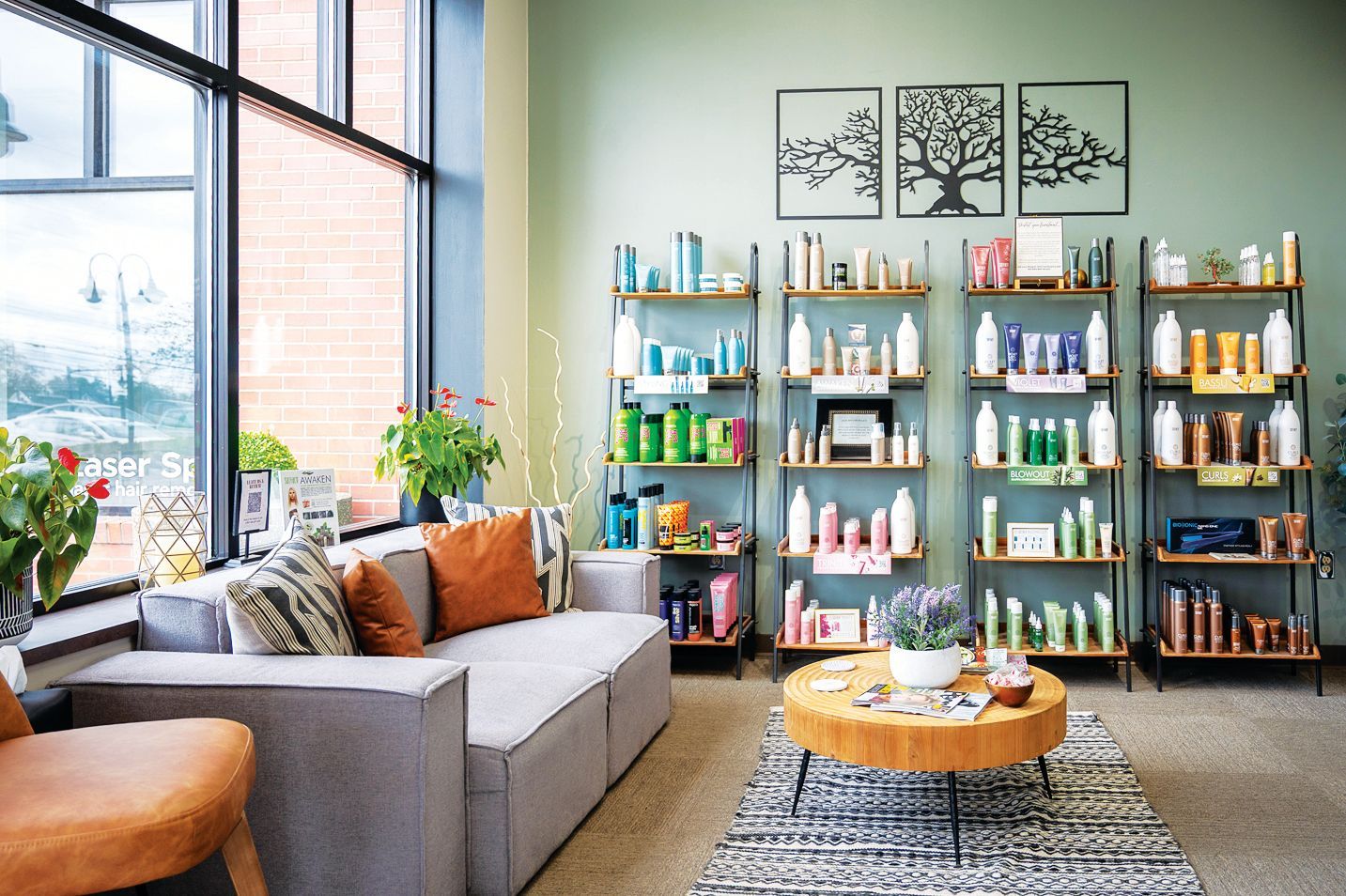 A brightly lit salon waiting area featuring a grey sofa, wooden coffee table, and tall shelves stocked with hair products.