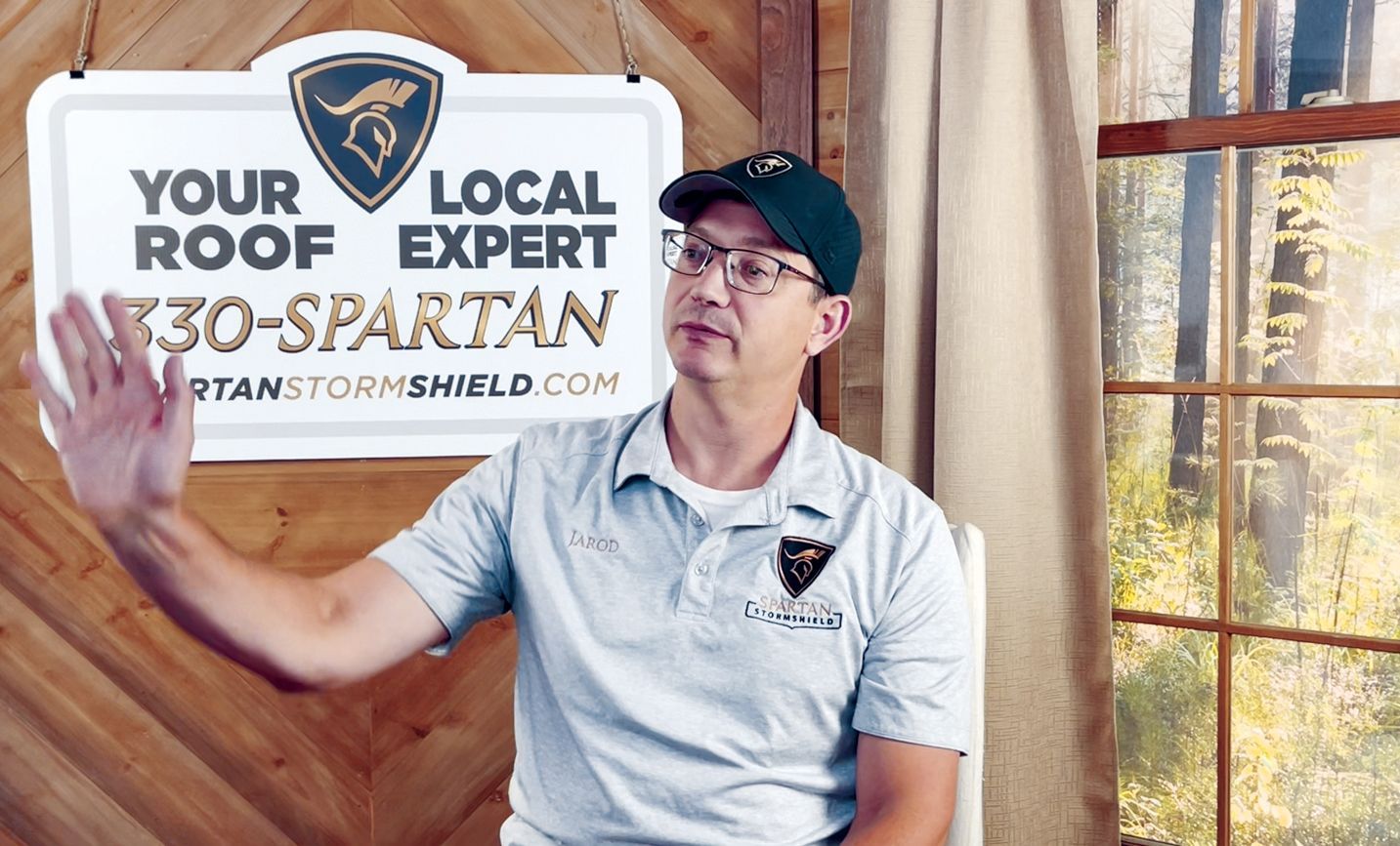 A man in a company polo and cap gestures while sitting in front of a sign for Spartan Storm Shield roofing.
