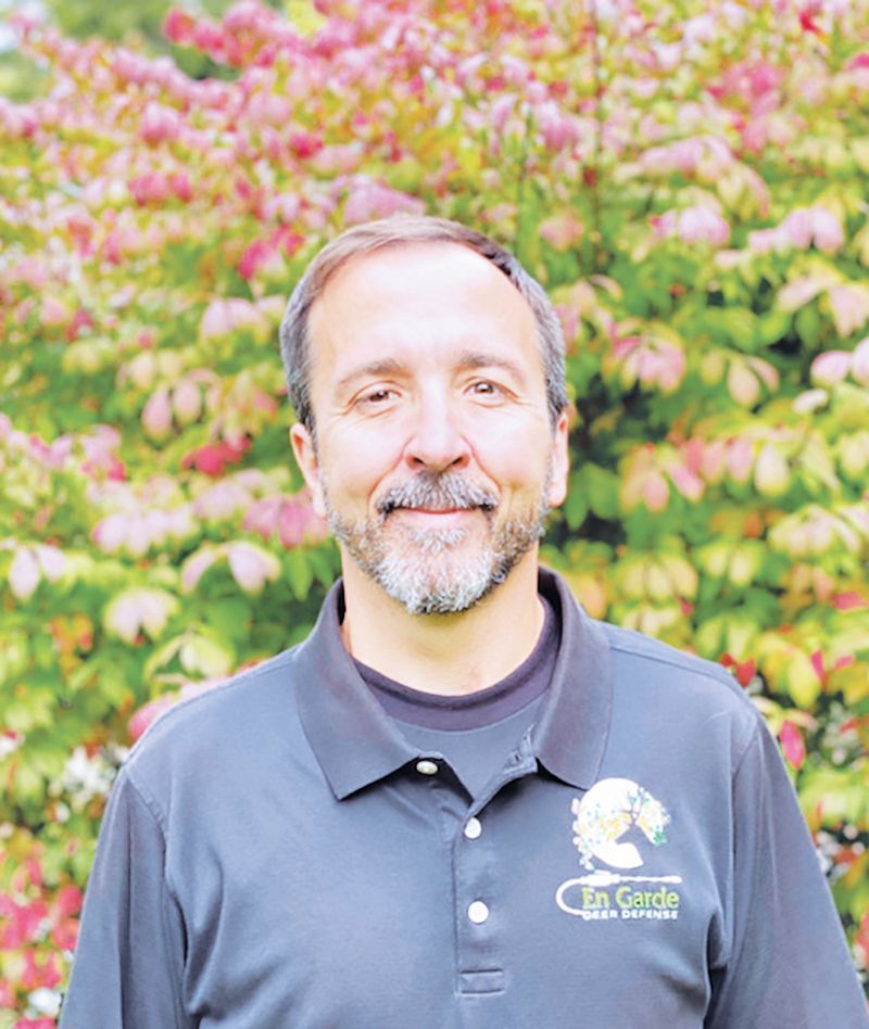 A man with a goatee wearing a gray polo shirt with an En Garde logo, standing in front of leafy bushes.