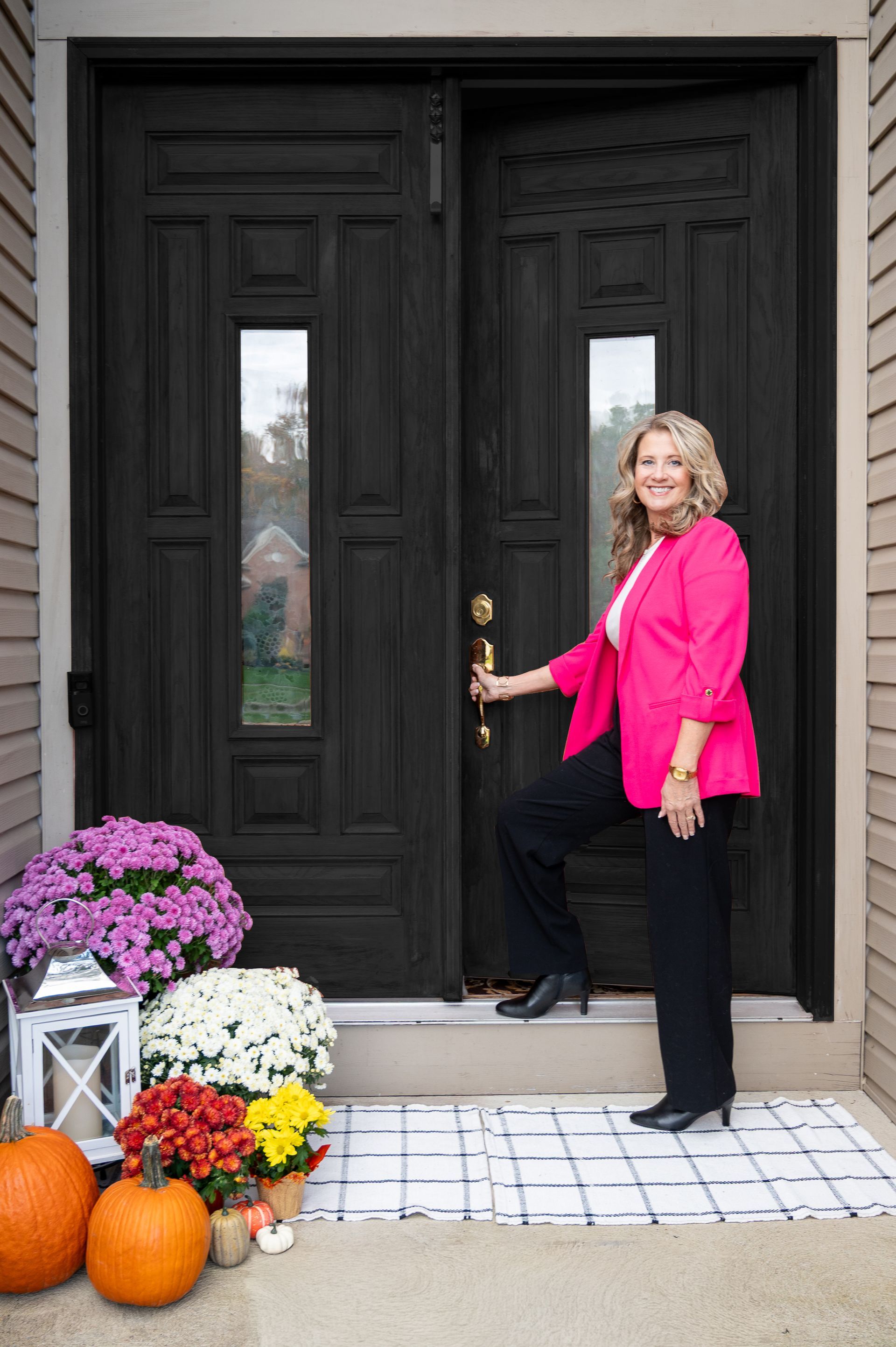 A smiling person in a bright pink blazer holds a black double door, surrounded by pumpkins and potted mums on a porch.