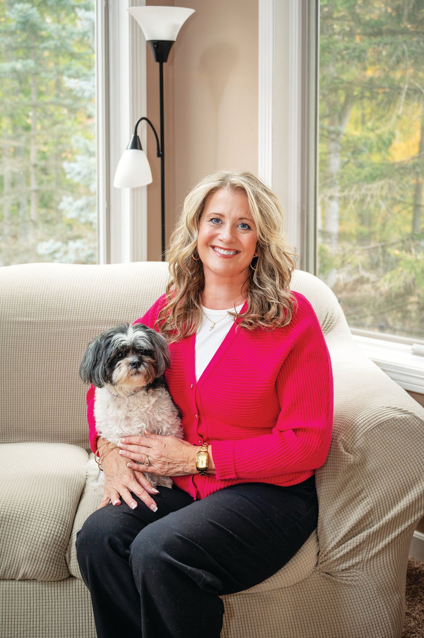 A smiling person sits on a beige sofa holding a small, black-and-white dog while wearing a bright red cardigan.