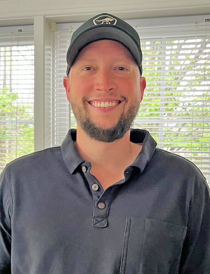 A smiling person with a dark beard and cap wearing a navy blue polo shirt, positioned in front of a window with blinds.