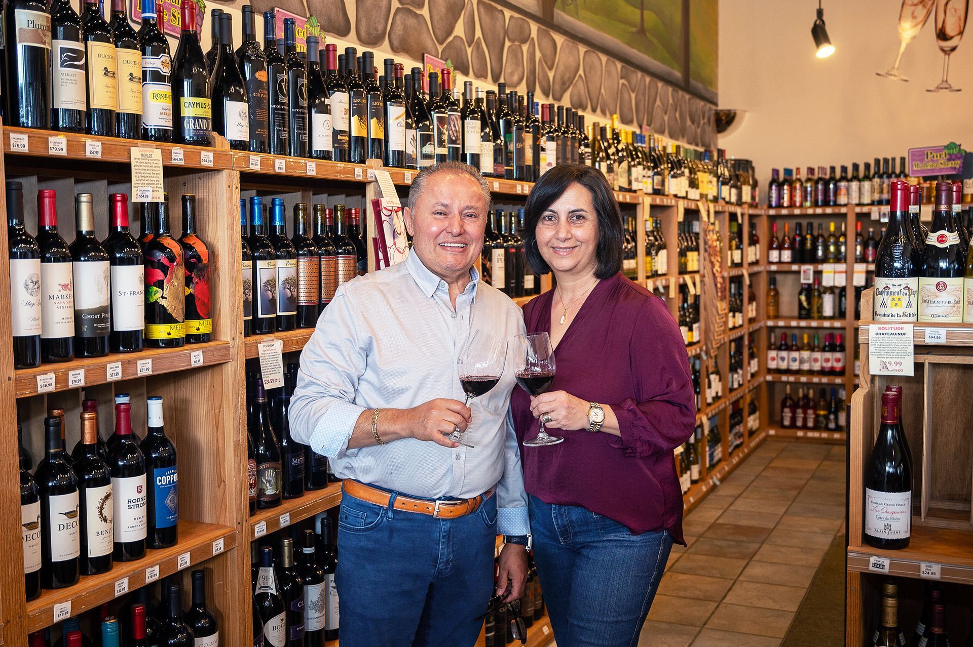 A smiling couple stands in a wine shop, each holding a glass of red wine, surrounded by shelves stocked with wine bottles.