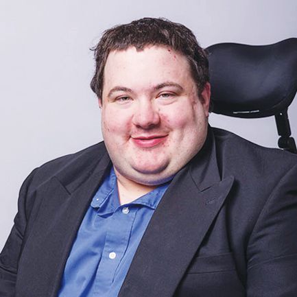 A smiling person in a blue collared shirt and black blazer sits in a wheelchair against a plain grey background.