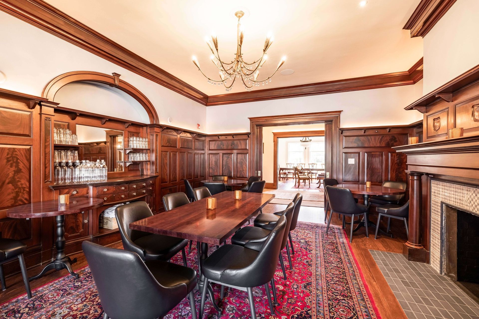 Elegant wood-paneled dining room with dark chairs, long tables, a crystal chandelier, and a large patterned rug.