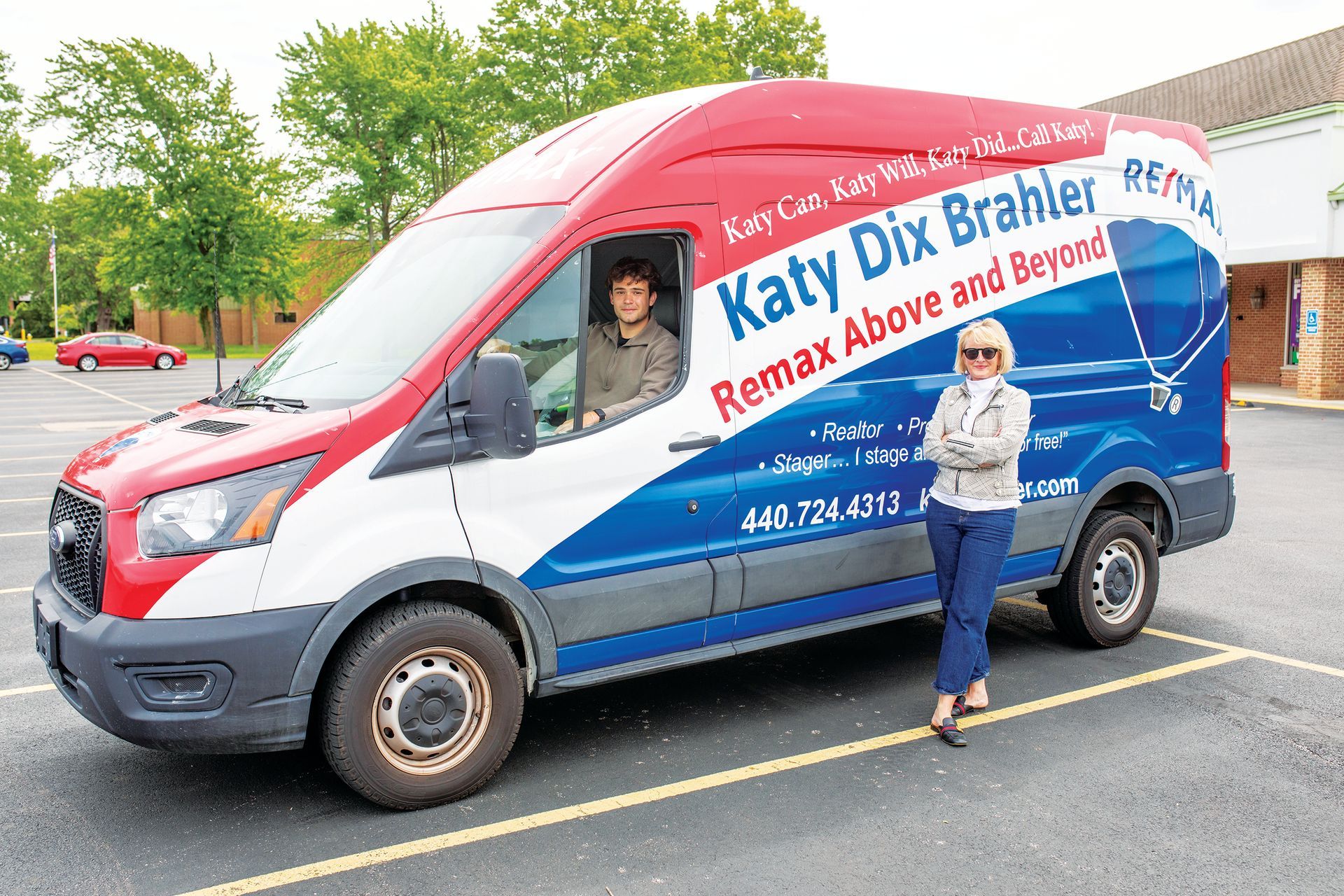 A person sits in the driver's seat of a branded RE/MAX van while another person stands beside it in a parking lot.