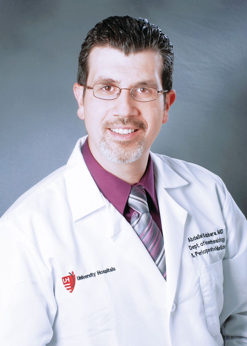 A smiling doctor wearing glasses, a white lab coat, and a purple shirt with a patterned tie against a gray background.