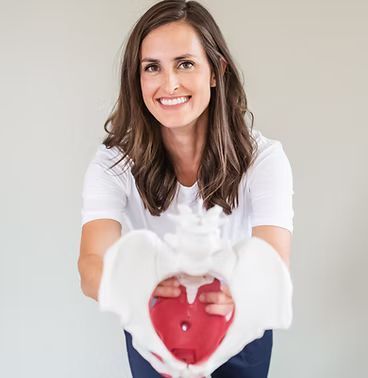 A person smiling while holding a model of a human pelvis toward the camera against a plain background.