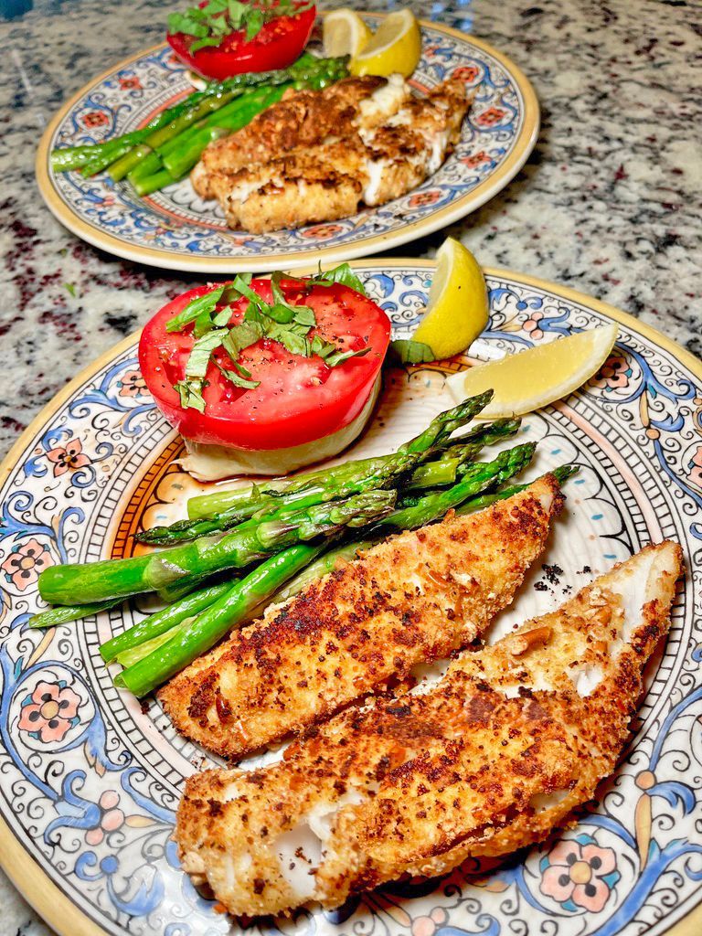Two plates each holding breaded fish, roasted asparagus, and a tomato slice topped with herbs, served with lemon wedges.