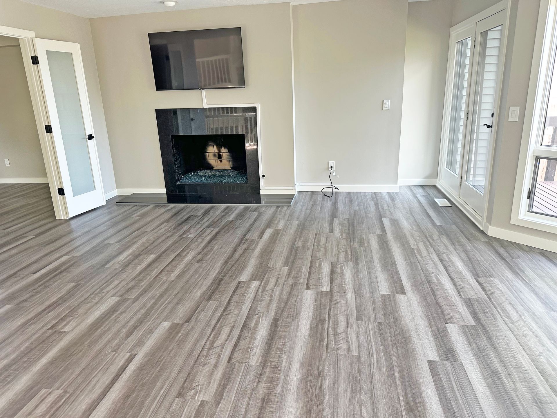 A bright room with grey wood-look flooring, a fireplace with a mounted television, and a white door and glass patio doors.