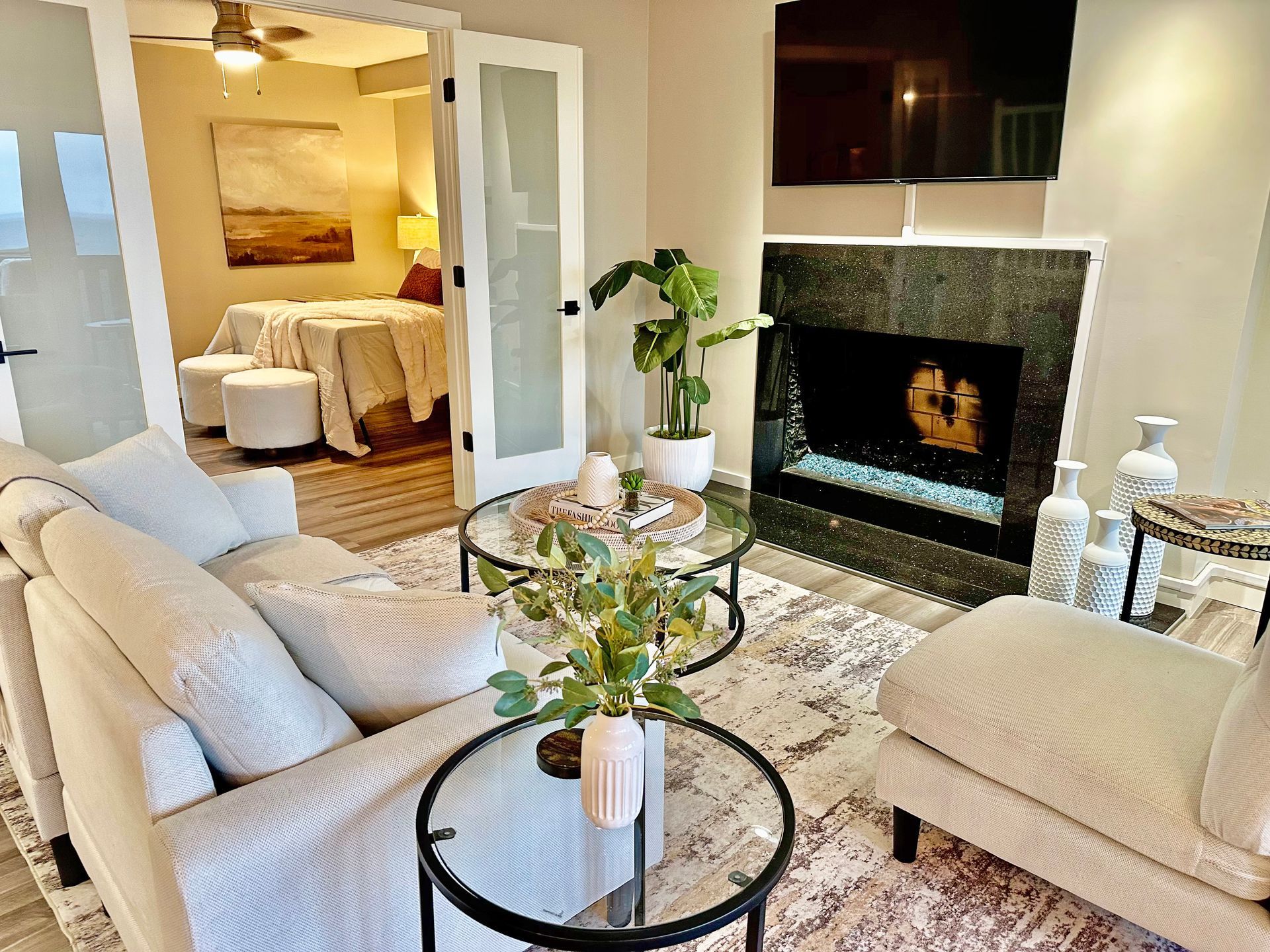 A neutral-toned living room features a fireplace, plush seating, glass-top coffee tables, and a view into a bedroom.