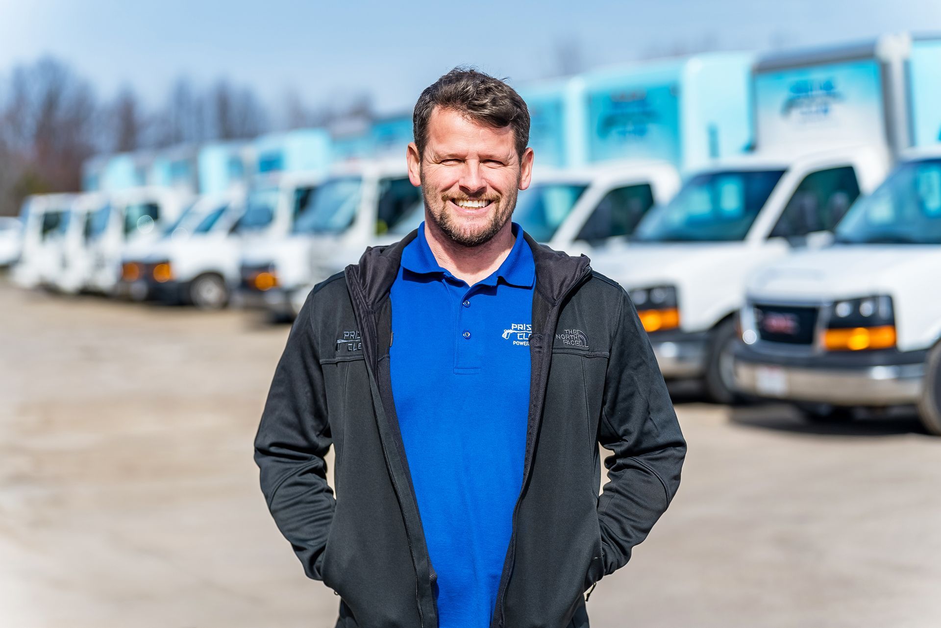 A smiling man in a blue polo and black hoodie stands in a parking lot in front of several white delivery trucks.