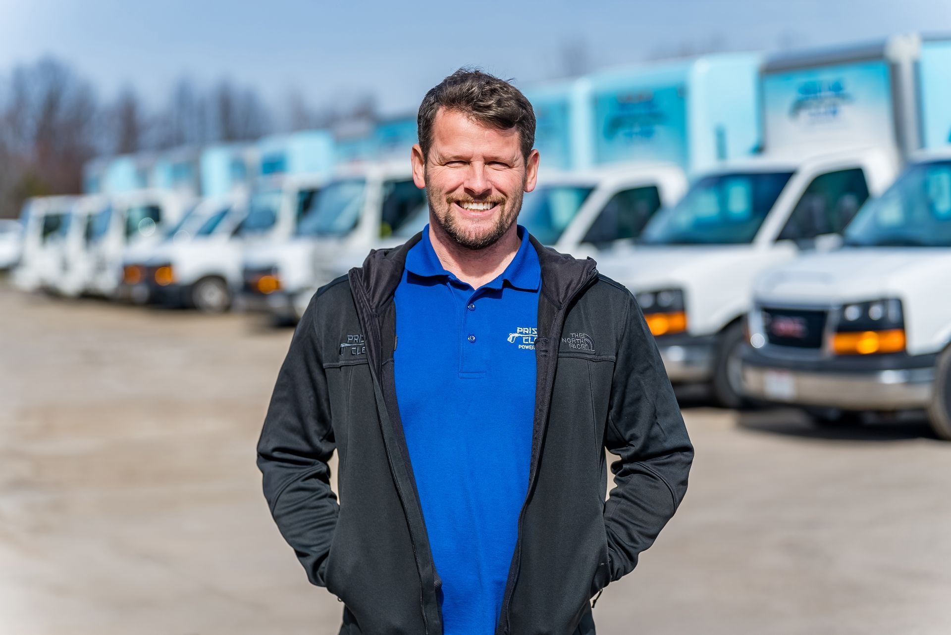 A smiling person in a blue polo shirt and black jacket stands in front of a row of parked white delivery trucks.