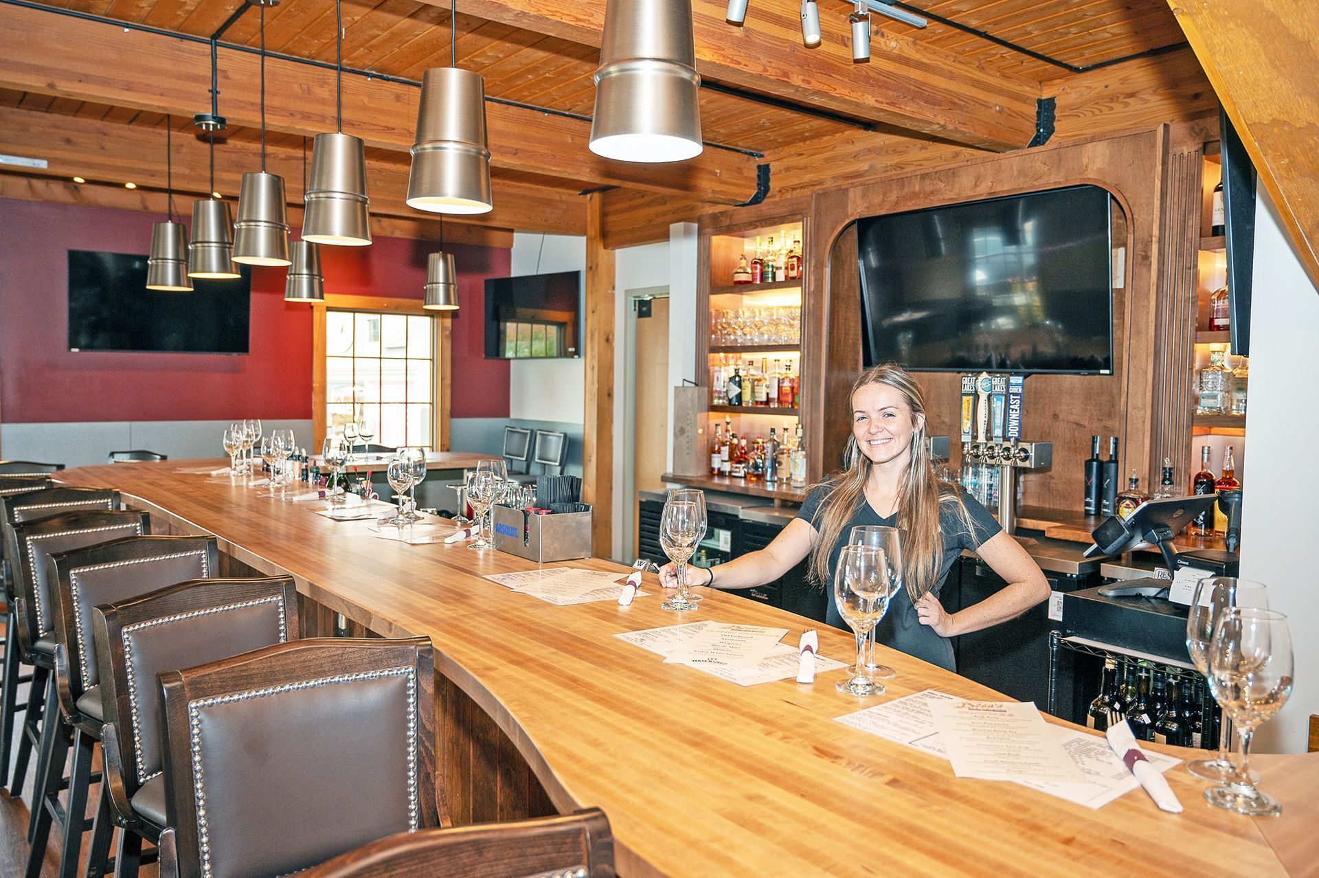 A smiling bartender stands behind a long wooden bar in a rustic, well-lit restaurant with leather chairs and hanging lamps.