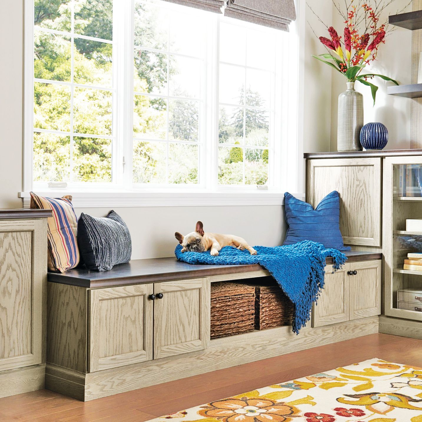 A French bulldog rests on a built-in window seat with storage cabinets, colorful cushions, and a blue throw blanket.