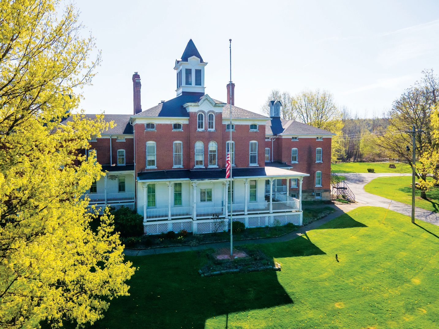 An aerial view of a historic red brick building with a central cupola and white porch, set on a bright green lawn.