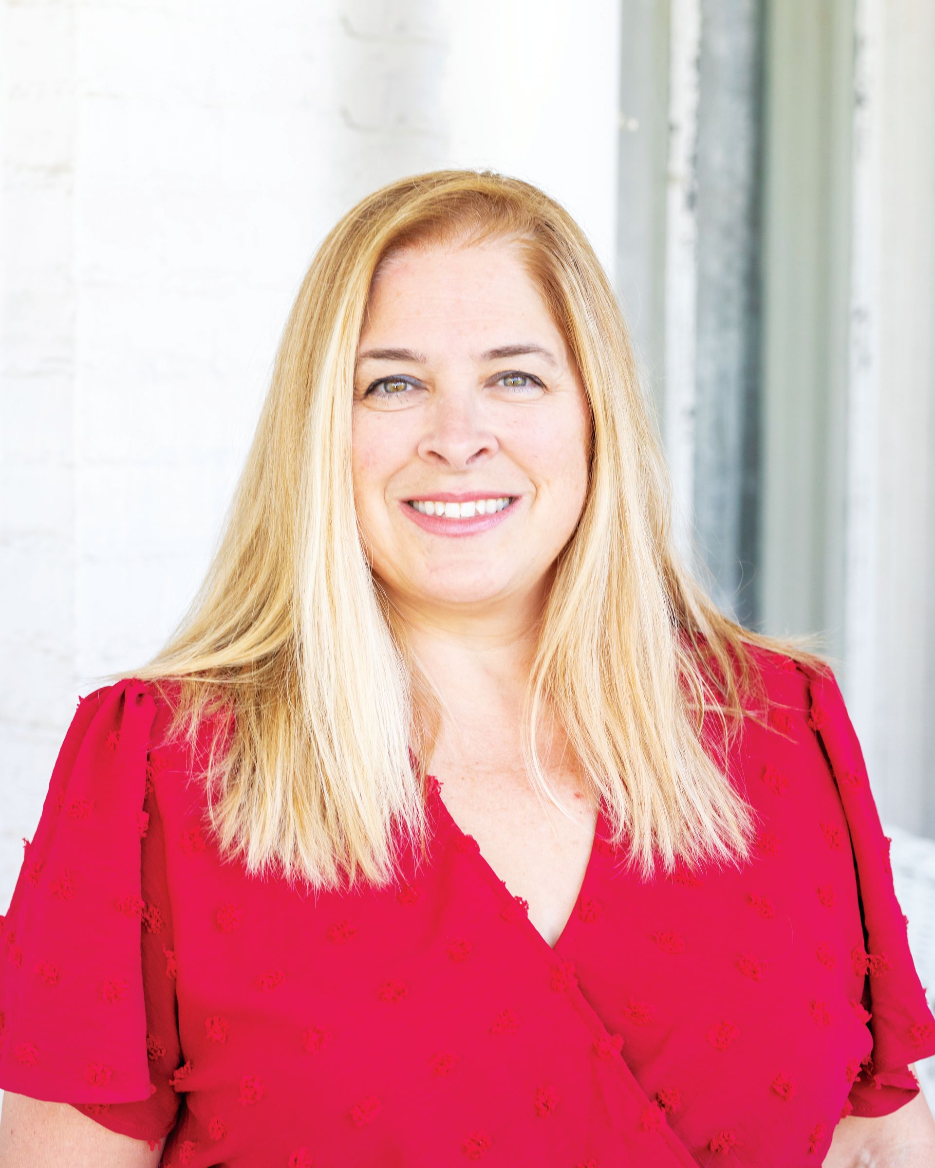 A person with shoulder-length blonde hair wearing a bright red top, smiling against a neutral, light background.