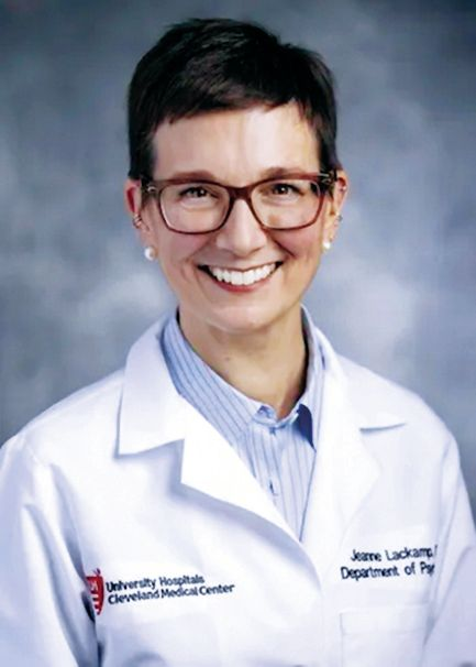 A professional portrait of a doctor wearing glasses and a white coat with a hospital logo, smiling against a blue background.