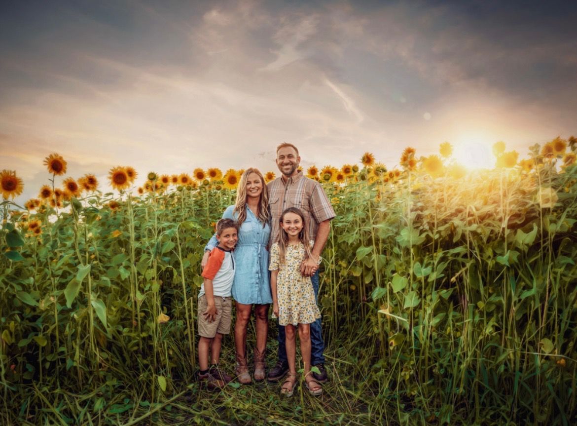 A family of four stands together in a large sunflower field during a warm, golden sunset.
