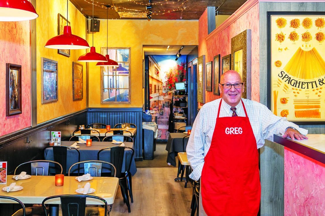 A smiling person in a red apron stands behind a counter in a warm-toned restaurant with tables and red pendant lights.