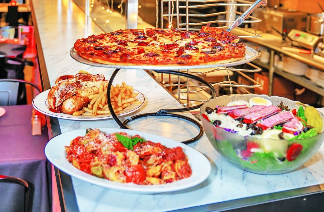 A spread of restaurant dishes featuring a pepperoni pizza, fries, chicken, a pasta dish, and a large side salad.