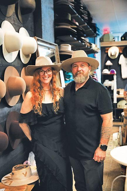 A woman and a bearded man wearing fedoras stand together in a hat shop filled with displays of hats.