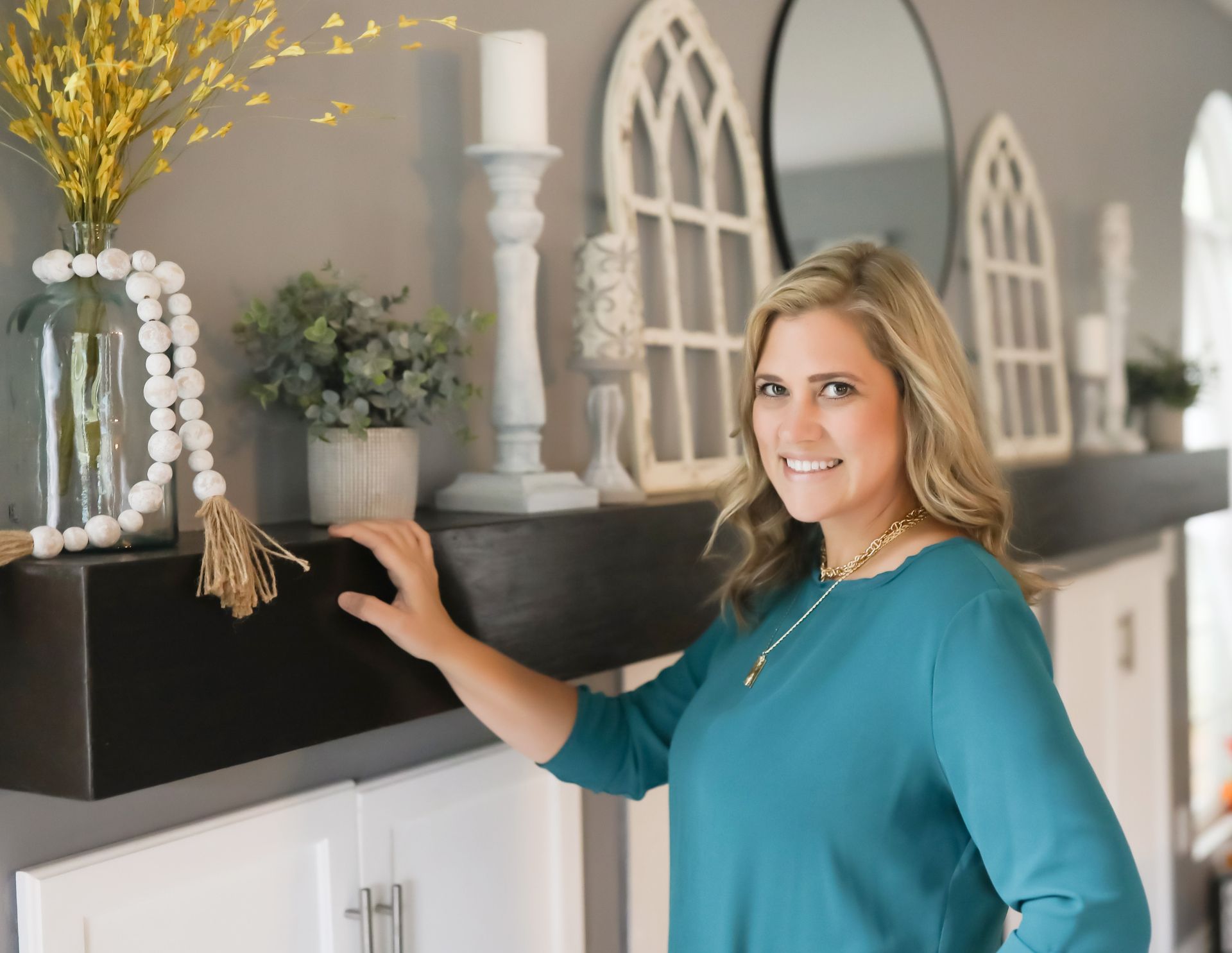 A smiling person in a teal shirt poses by a dark wooden mantel decorated with a vase, beads, greenery, and wall art.