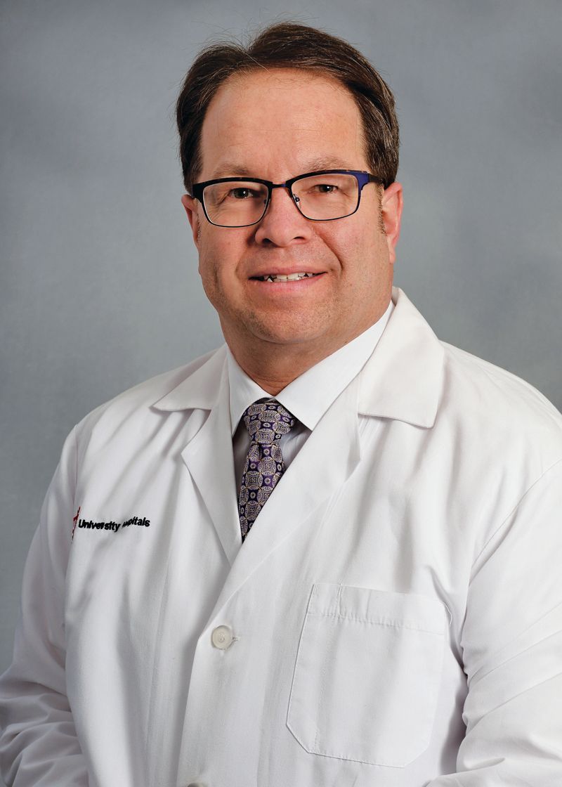 A person wearing a white lab coat, dress shirt, and patterned tie, posing against a neutral gray background.