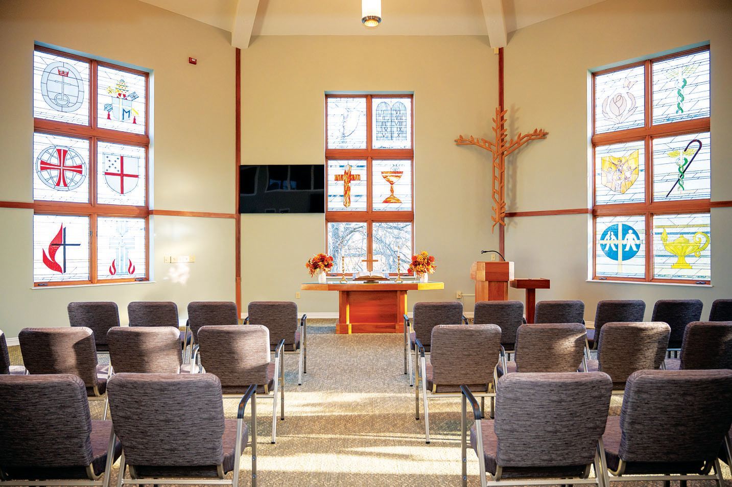 Rows of chairs face a central altar and wooden cross in a chapel with stained glass windows on the side walls.