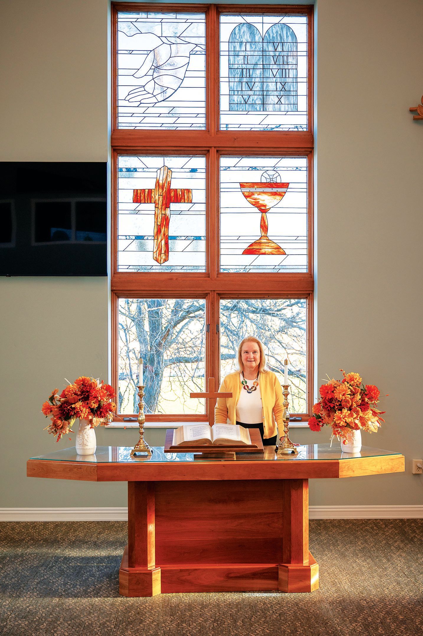 A person stands behind a wooden altar in front of a stained-glass window featuring a cross and a chalice.