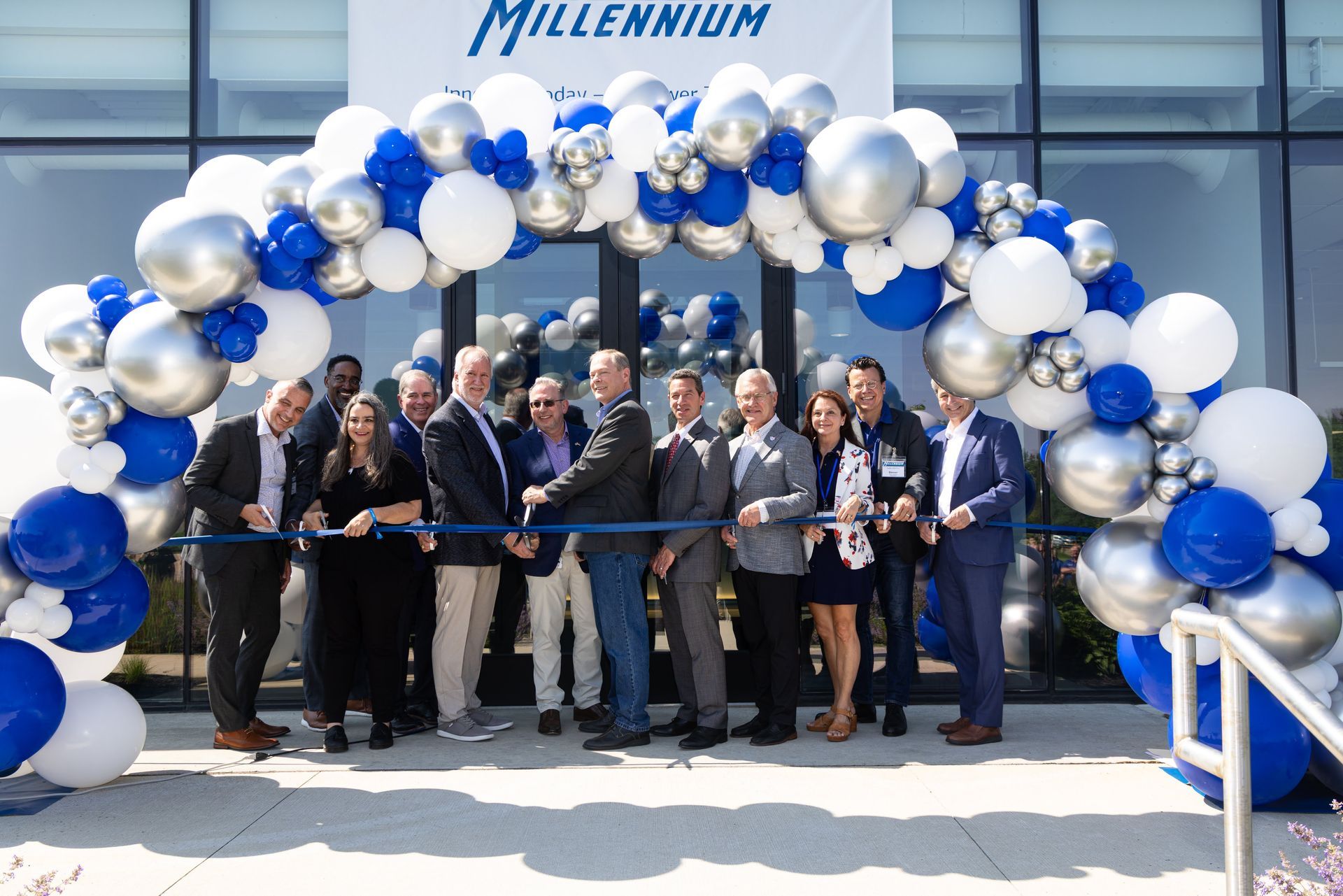 A group of people standing in front of a building during a ribbon-cutting ceremony under a blue, silver, and white arch.