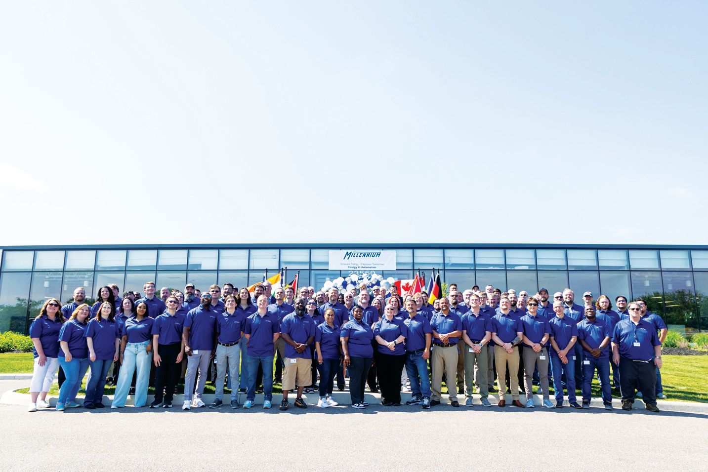 A large group of people in blue shirts stand in front of a modern glass office building on a sunny day.