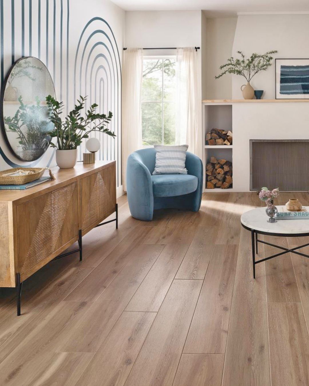 A bright living room with wood floors, a light blue armchair, a wooden console table, a round mirror, and a fireplace.