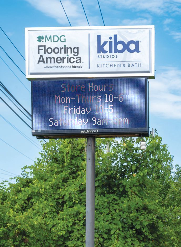 A freestanding outdoor business sign for MDG Flooring America and Kiba Studios against a blue sky with green trees below.