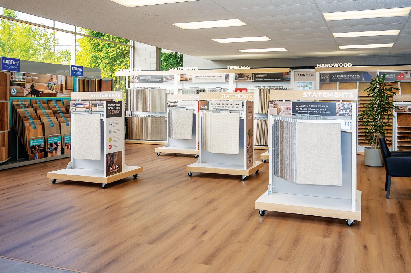 A bright showroom featuring multiple floor-standing displays of carpets and wood flooring samples on a wooden floor.